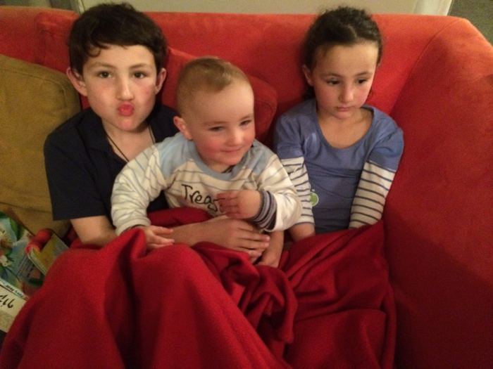 Three children under a red blanket on a sofa